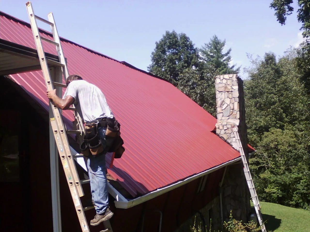 Roofer on a ladder beside a red metal roof and stone chimney in Clay Center